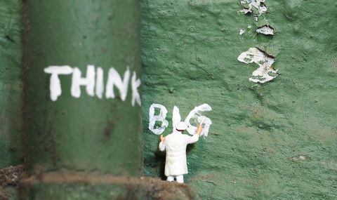 Miniaturmännchen vor grünem Hintergrund, das Think Big an die Wand schreibt